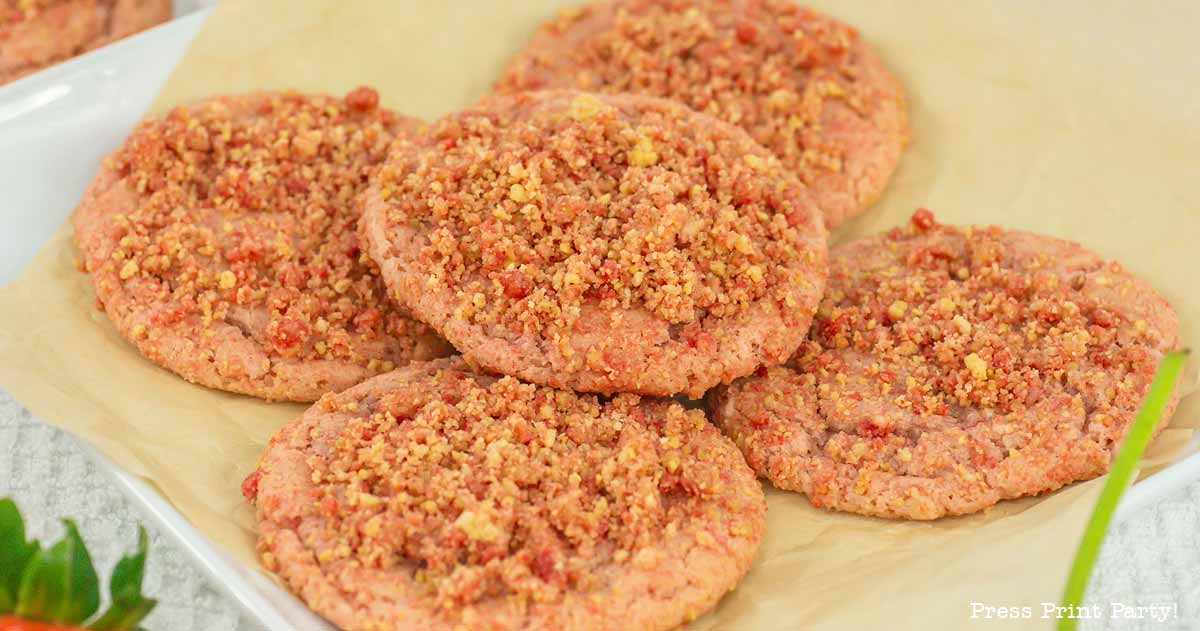 A plate of cookies topped with a crumbly mixture, placed on parchment paper. The cookies have a light pink hue, suggesting a fruity or flavored variation. They are arranged in a slightly overlapping pattern.