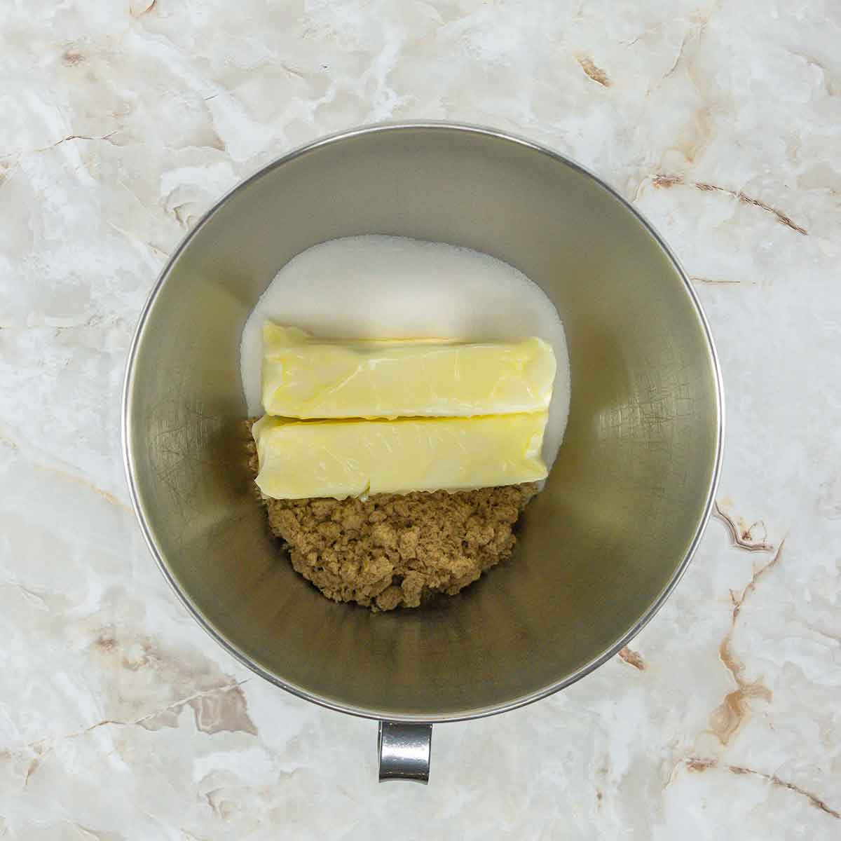 A metal mixing bowl on a marble surface containing two sticks of butter, granulated sugar, and brown sugar. The ingredients are arranged side by side, ready for mixing.