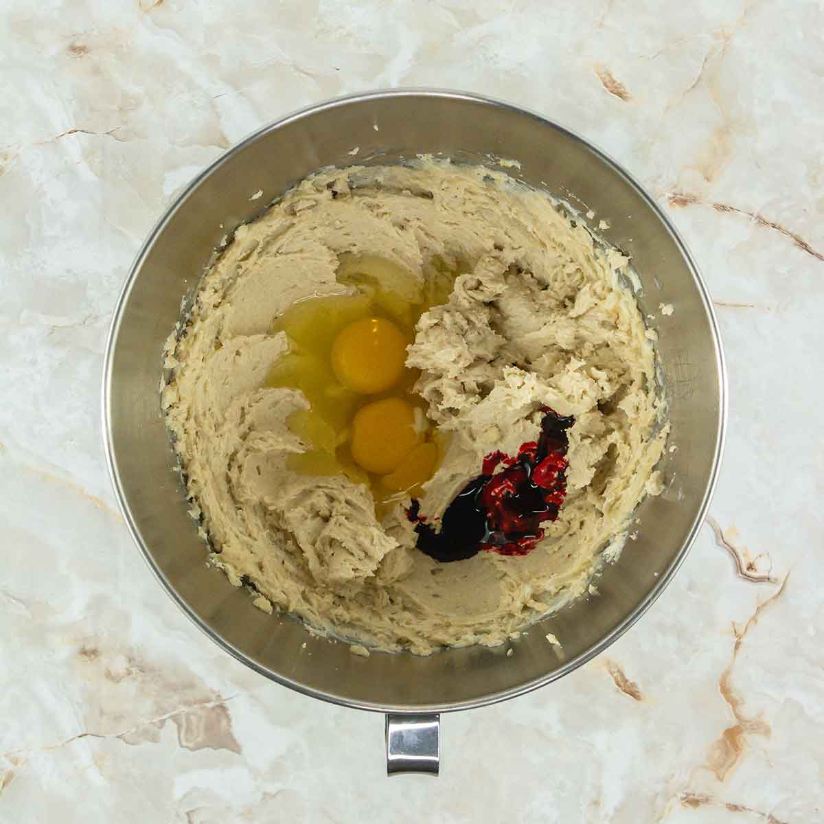 A metal mixing bowl contains creamy batter with two cracked eggs and red food coloring on a marbled countertop.