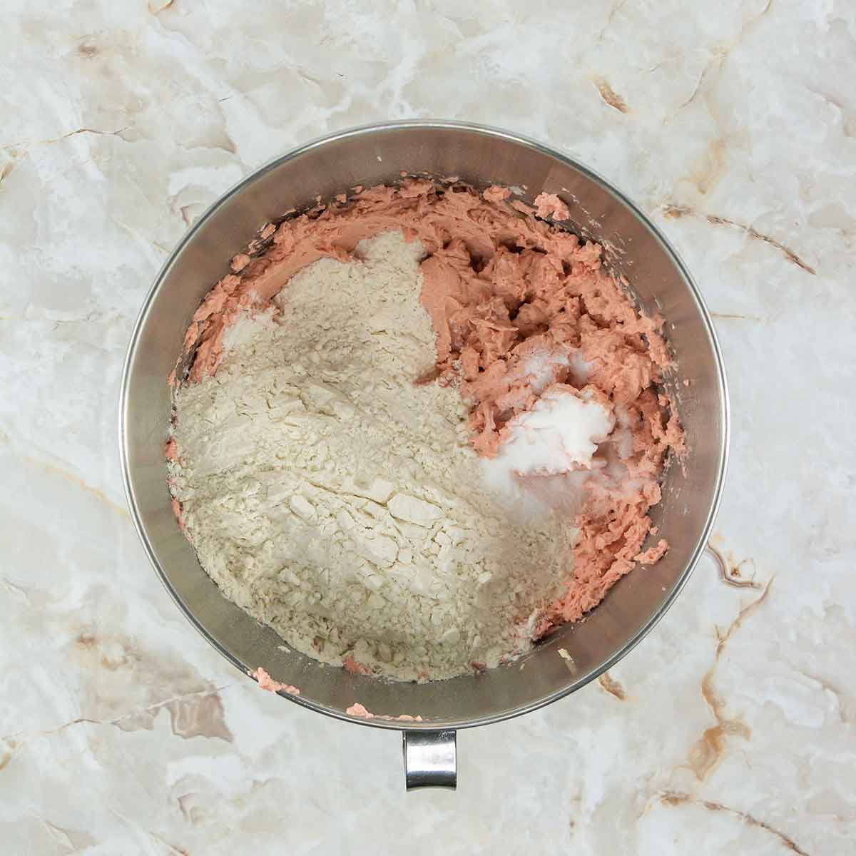 A mixing bowl containing pink dough partially mixed with white flour and a small amount of a white substance, likely a type of fat or shortening, on a marble countertop.