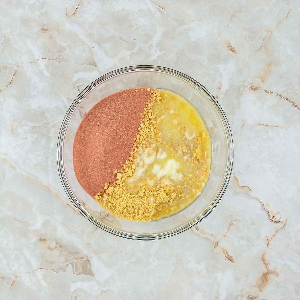 A glass bowl containing a mixture of red powder, crushed biscuits, and a yellow liquid on one side, all set on a light marble surface.