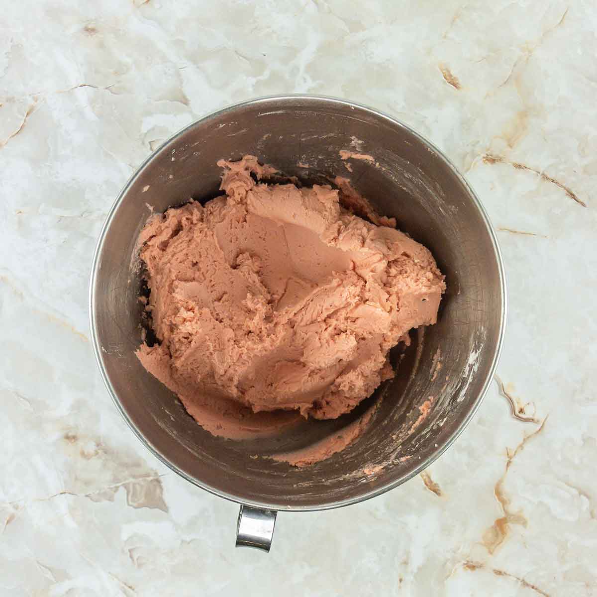 A metal mixing bowl with light pink dough sits on a marble countertop.