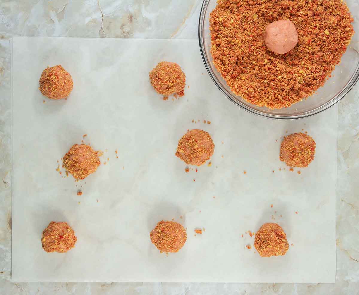 Overhead view of eight coated, round snack balls on parchment paper. A glass bowl containing one unrolled ball and a coarse, reddish seasoning mix is visible on the side. They are set on a marble countertop.