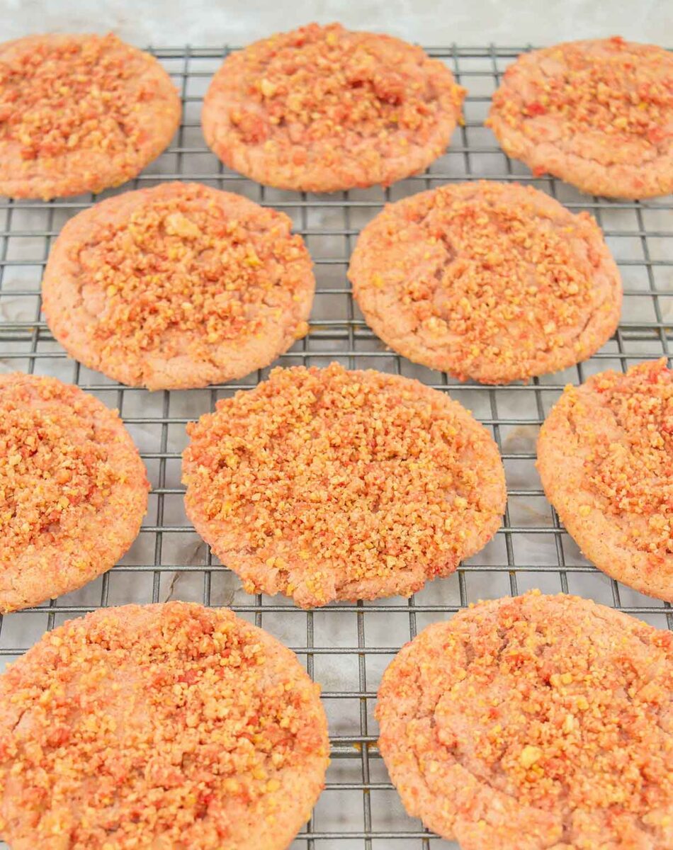 Nine strawberry crumb cookies are cooling on a wire rack. Each cookie is topped with a crumbly mixture, giving them a textured appearance. They are arranged in three rows on a light-colored surface.