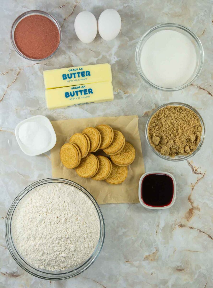 Top-down view of baking ingredients on a marble surface: two eggs, two butter sticks, sugar in a bowl, brown sugar in a bowl, golden sandwich cookies on parchment, a bowl of flour, cocoa powder, vanilla extract, and a small bowl of salt.