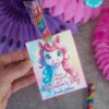 A hand displays the "template (Copy) (Copy)" Valentine's Day card. It features a unicorn with rainbow hair and the message, "Have a magical Valentine's Day," paired with an attached rainbow lollipop. Pink and purple decorations adorn the background.