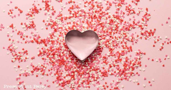 A pink heart-shaped cookie cutter is adorned with scattered red, pink, and white heart-shaped sprinkles on a pink background—perfect for inspiring Valentine's Day party ideas.