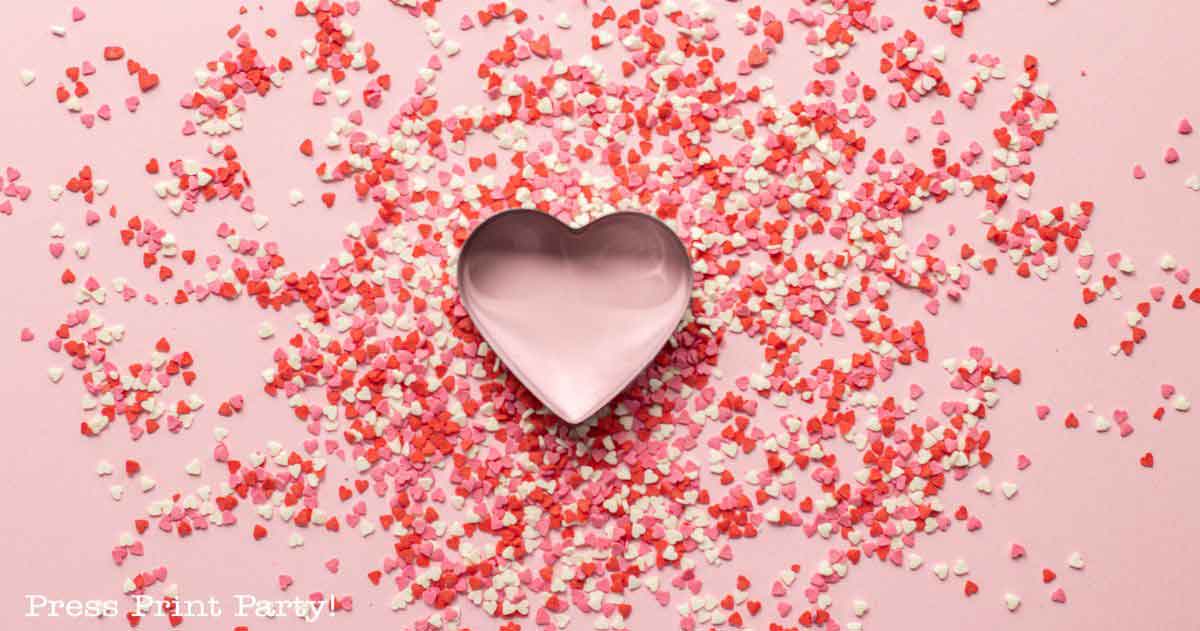 A pink heart-shaped cookie cutter is adorned with scattered red, pink, and white heart-shaped sprinkles on a pink background—perfect for inspiring Valentine's Day party ideas.
