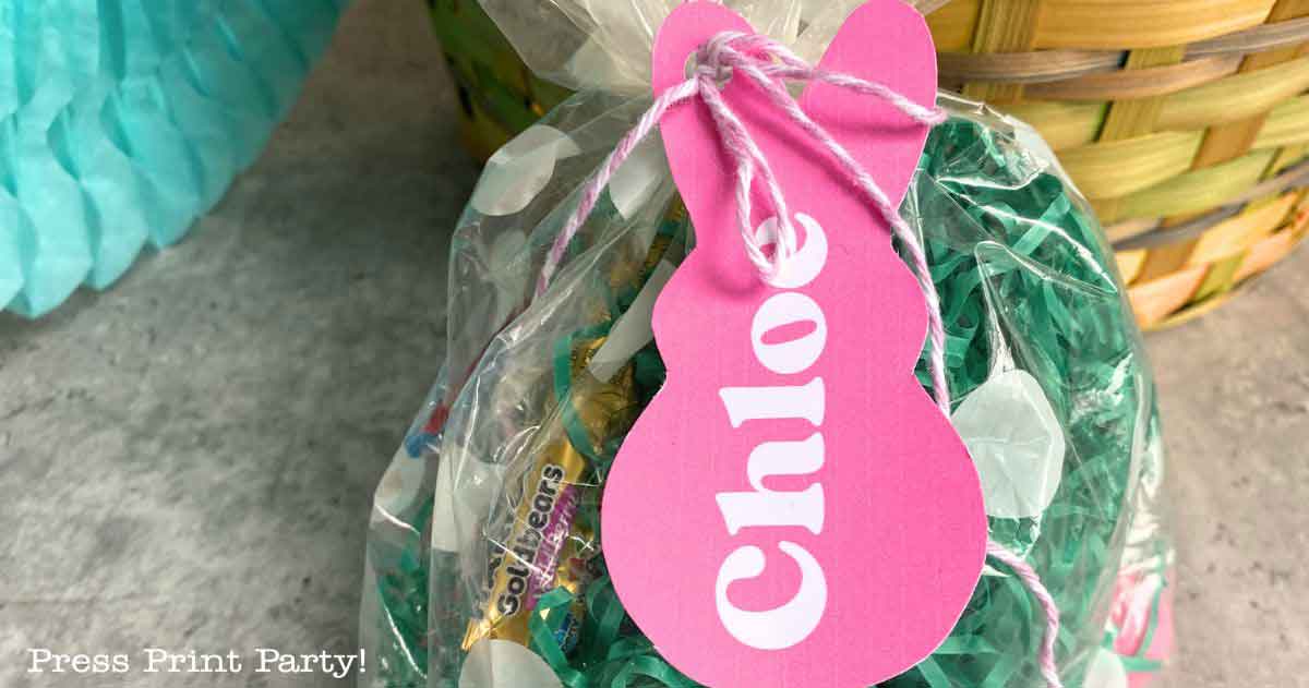 A clear plastic bag filled with Easter candy and green shredded paper is tied with pink and white string. An Easter basket tag in the shape of a pink paper bunny with the name "Chloe" is attached. In the background, a wicker basket and blue decoration complete the festive scene.