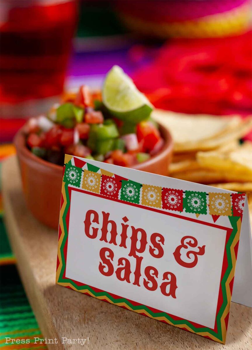 Colorful table setting with a card labeled "Chips & Salsa" in festive font. A bowl of salsa with a lime wedge is in the background, alongside a stack of tortilla chips on a wooden board. Bright colors and decorative patterns enhance the festive atmosphere.