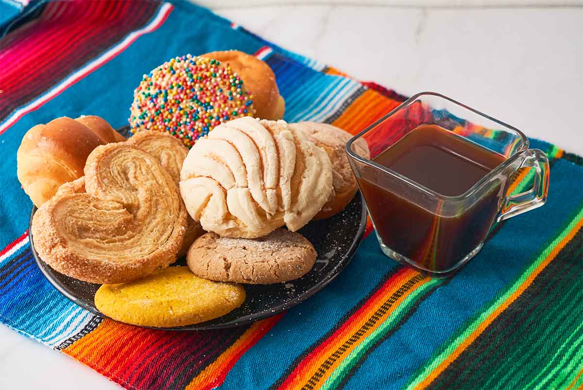 A variety of Mexican pastries, including a concha, palmera, and cookies, are arranged on a black plate. A glass cup of coffee is next to the plate, all set on a colorful striped fabric.