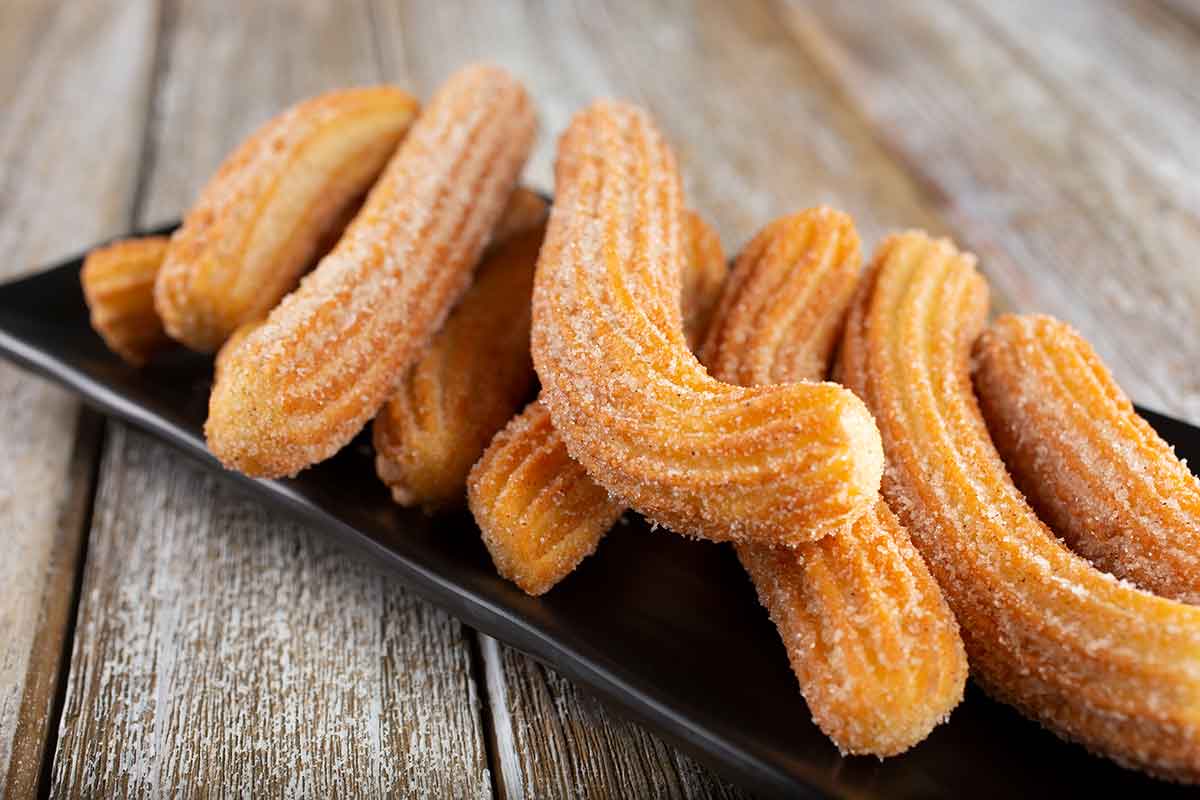 A black rectangular plate holds several golden-brown churros coated in sugar. The churros are arranged on a wooden table, their ridged surfaces and twisted shapes making a visually appealing display.