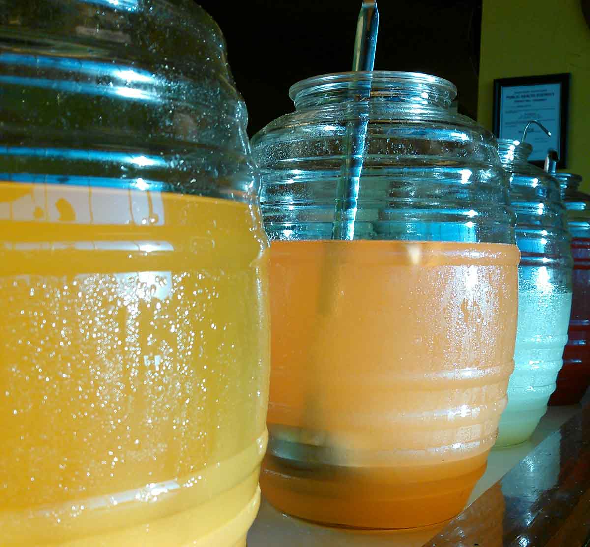 Large clear containers filled with various colorful beverages, including orange and yellow drinks, are lined up on a counter. Each container has a ladle. The containers are covered in condensation, suggesting they are cold.