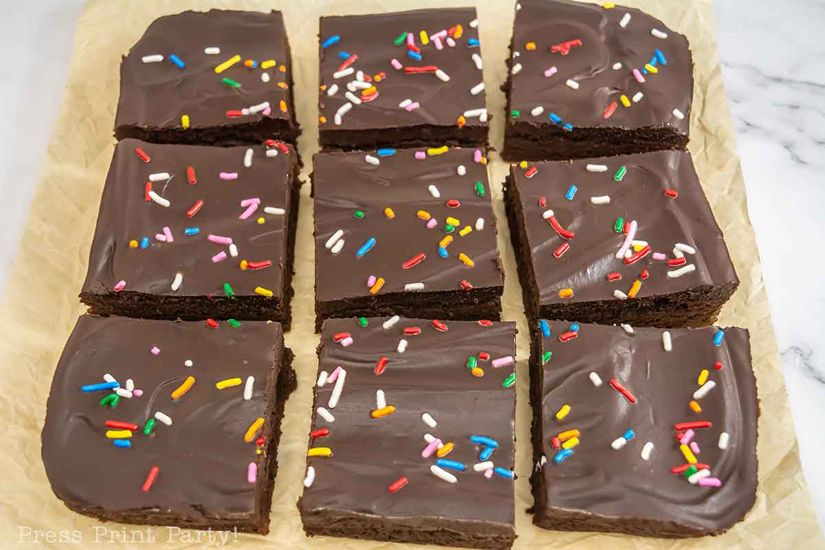 Nine chocolate frosted brownies with colorful sprinkles are arranged in a grid on parchment paper.