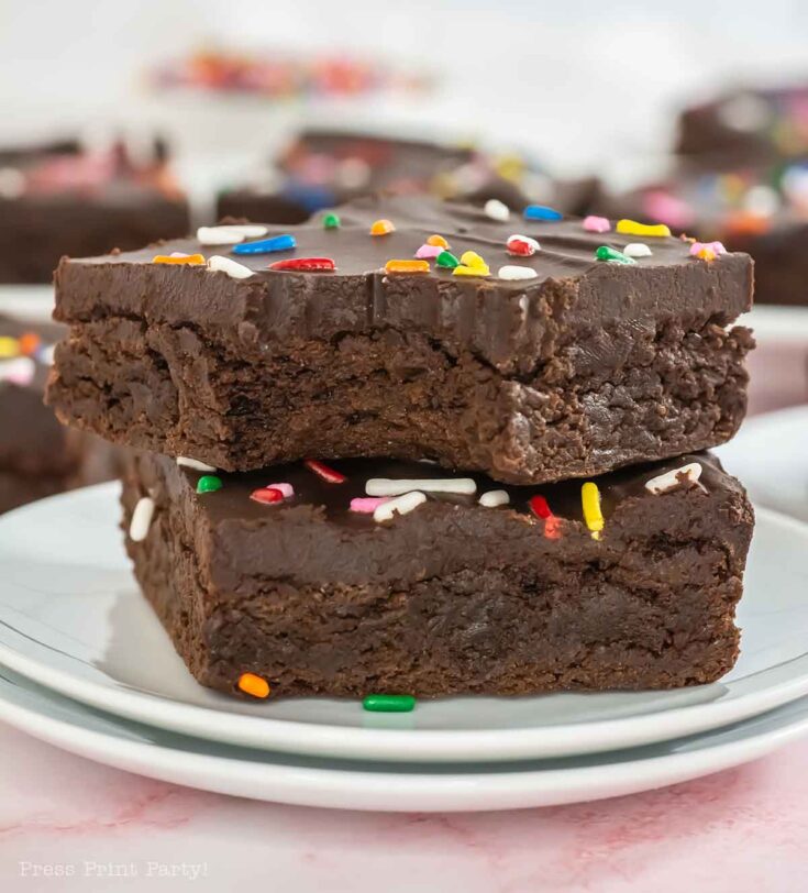 Two chocolate brownies topped with colorful sprinkles are stacked on a plate. The rich texture is visible, and the background shows more brownies slightly out of focus.