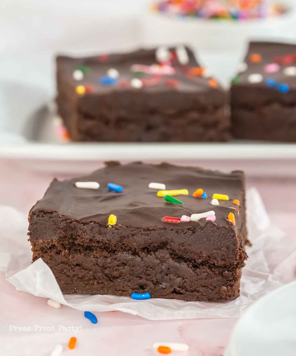 A close-up of a chocolate brownie decorated with colorful sprinkles on top. It rests on a piece of white parchment paper, with more brownies blurred in the background.
