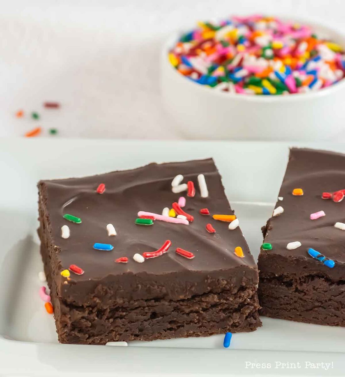 Two chocolate brownies topped with chocolate icing and colorful sprinkles are on a white plate. A bowl filled with more sprinkles is in the background.