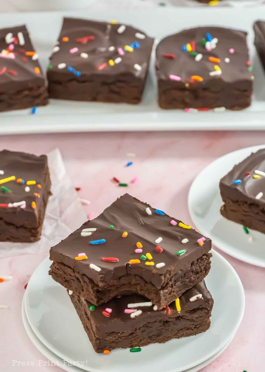 Chocolate frosted brownies with colorful sprinkles are stacked on white plates. More brownies are displayed in the background on a serving tray. The surface beneath is light pink with scattered sprinkles.