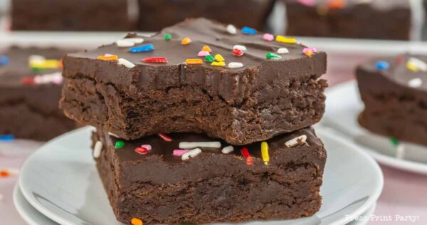 Two chocolate brownies topped with colorful sprinkles are stacked on a white plate. The brownie on top has a bite taken out of it, revealing a dense, rich texture.