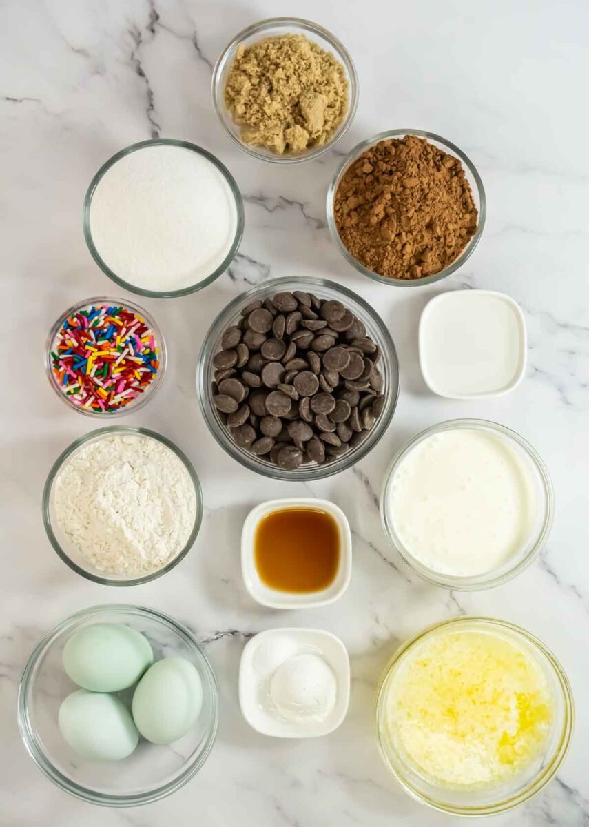 A collection of baking ingredients on a marble surface, including bowls of sugar, brown sugar, cocoa powder, chocolate chips, flour, rainbow sprinkles, vanilla extract, eggs, butter, cream, and baking soda.