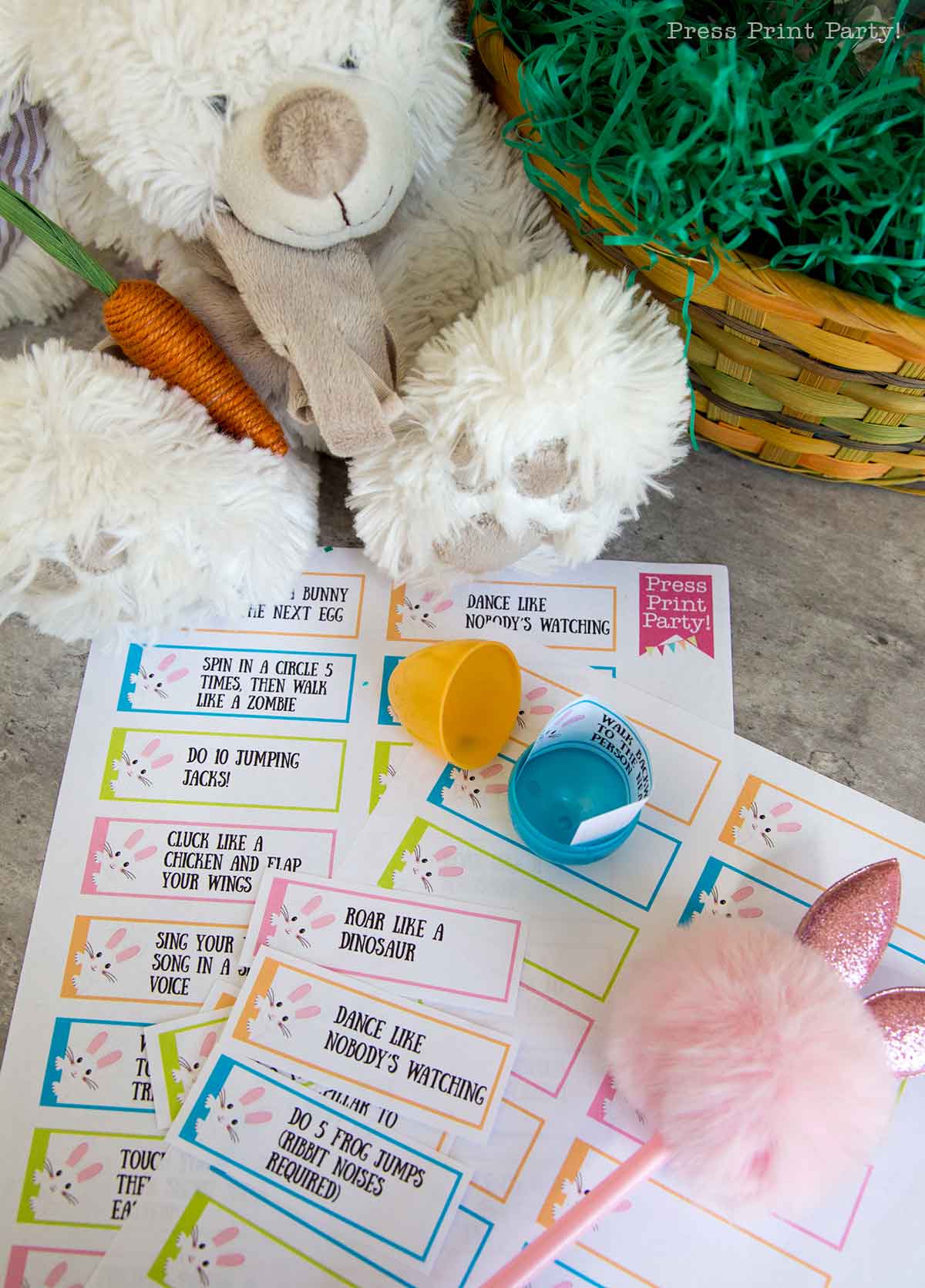 A stuffed white bear holds a plush carrot beside a basket with green shredded paper, perfect for an Easter egg hunt. In front are colorful activity cards with playful challenges, an open plastic egg, a pink pom-pom pen, and papers decorated with animals and text.