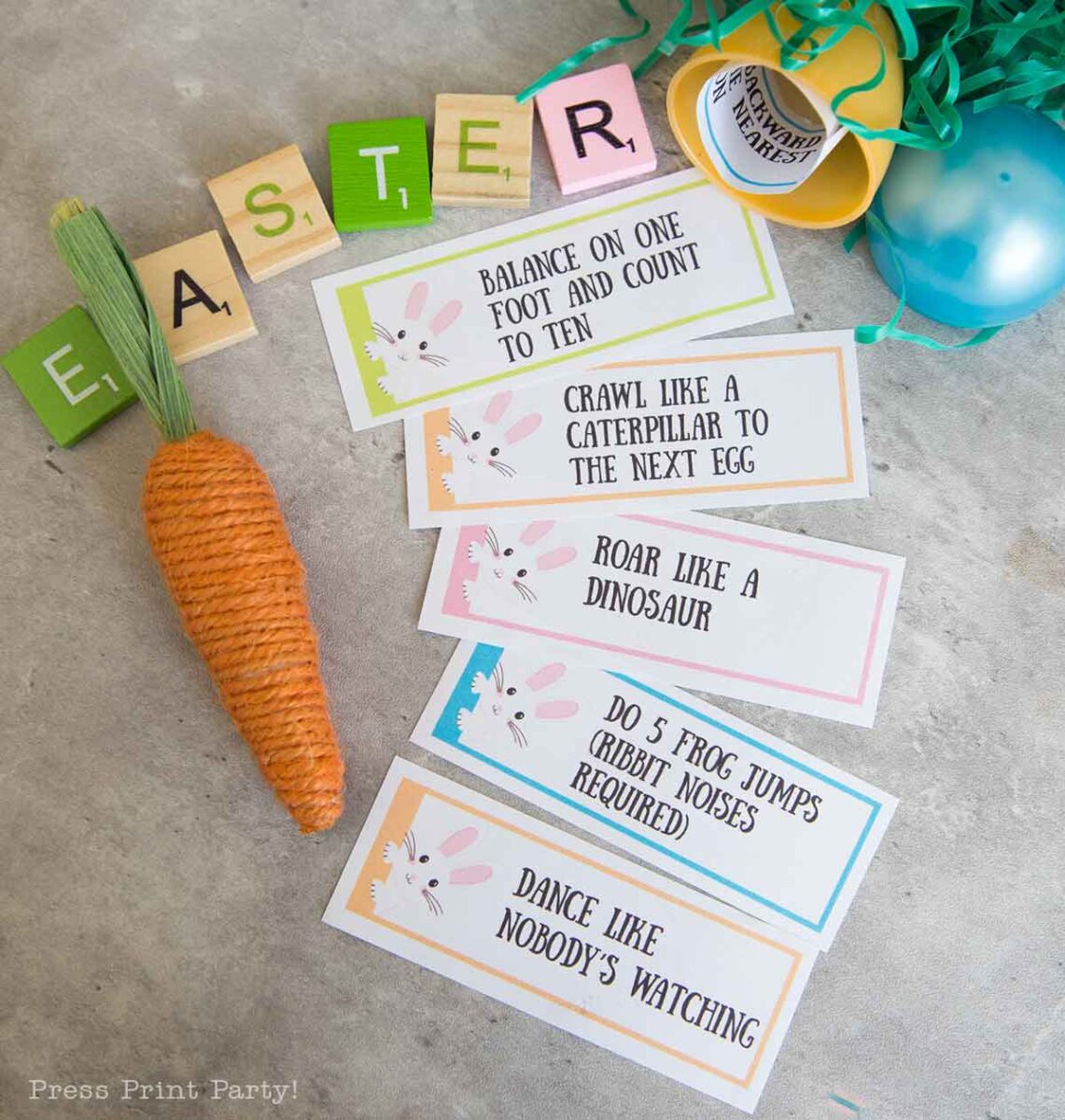 Easter-themed party game cards are arranged on a table alongside a decorative carrot and colorful letter blocks spelling "Easter." These Easter egg hunt activity cards feature playful actions like balancing, crawling, roaring, jumping, and dancing.