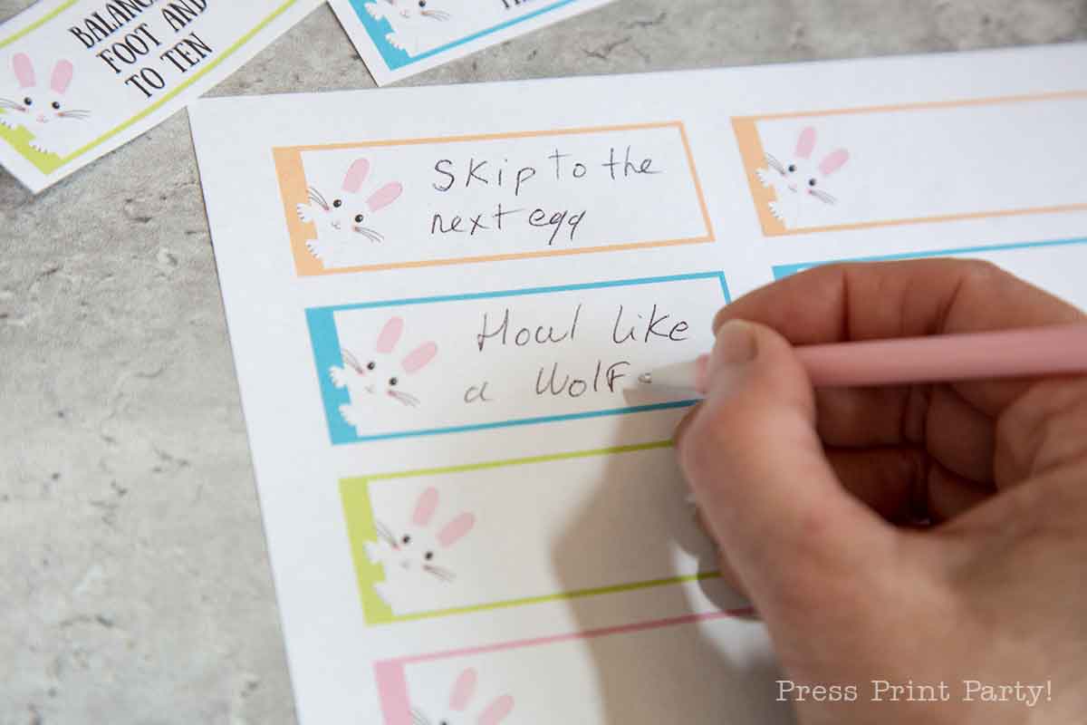A hand holds a pink pen, jotting "Skip to the next egg" and "Howl like a wolf" on pastel-colored easter egg hunt activity cards. Each card displays a charming bunny illustration, scattered across a gray surface.