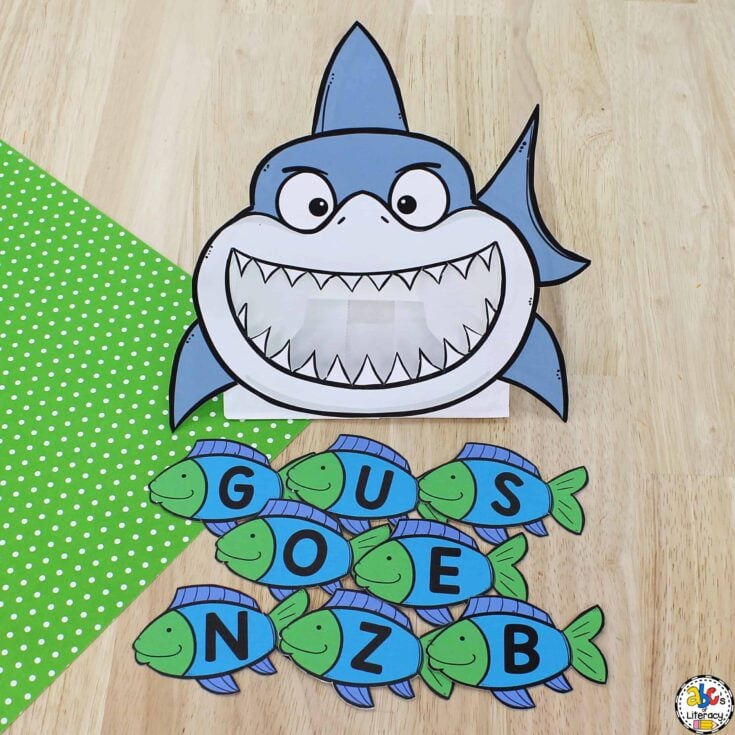 A paper shark with an open mouth is above seven paper fish, each with a bold black letter (G, U, S, O, E, N, Z, B). The items are on a wooden surface with a green polka dot cloth on the left.