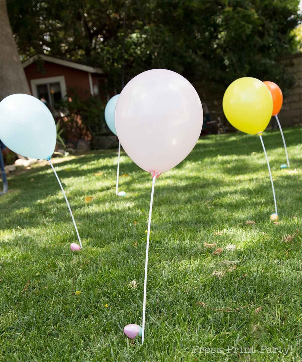Colorful balloons attached to white sticks stand upright on a grassy lawn, hinting at festive Easter Egg Scavenger Hunt ideas. A small shed and trees provide a charming backdrop for the balloons in shades of white, blue, yellow, and orange.