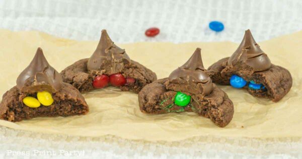 Four chocolate cookies topped with chocolate kisses are shown on parchment paper. Each cookie is cut in half, revealing colorful candy-coated chocolates inside: yellow, red, green, and blue.