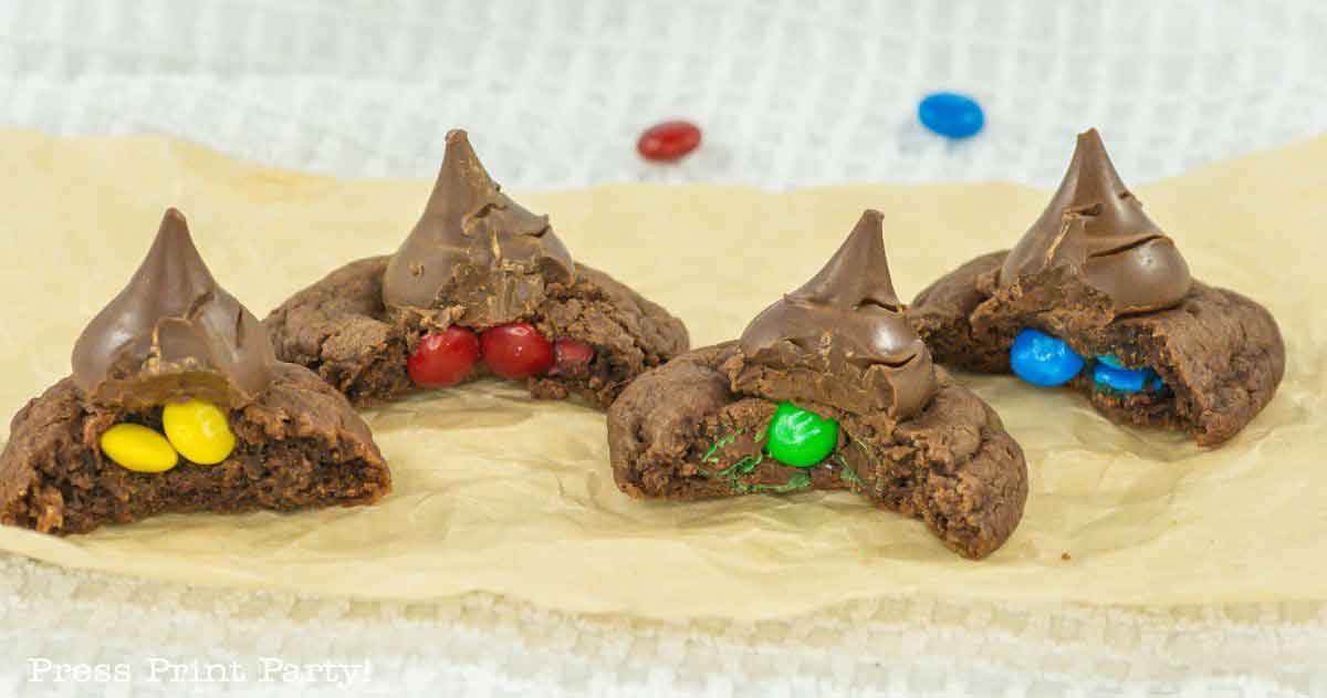 Four chocolate cookies topped with chocolate kisses are shown on parchment paper. Each cookie is cut in half, revealing colorful candy-coated chocolates inside: yellow, red, green, and blue.