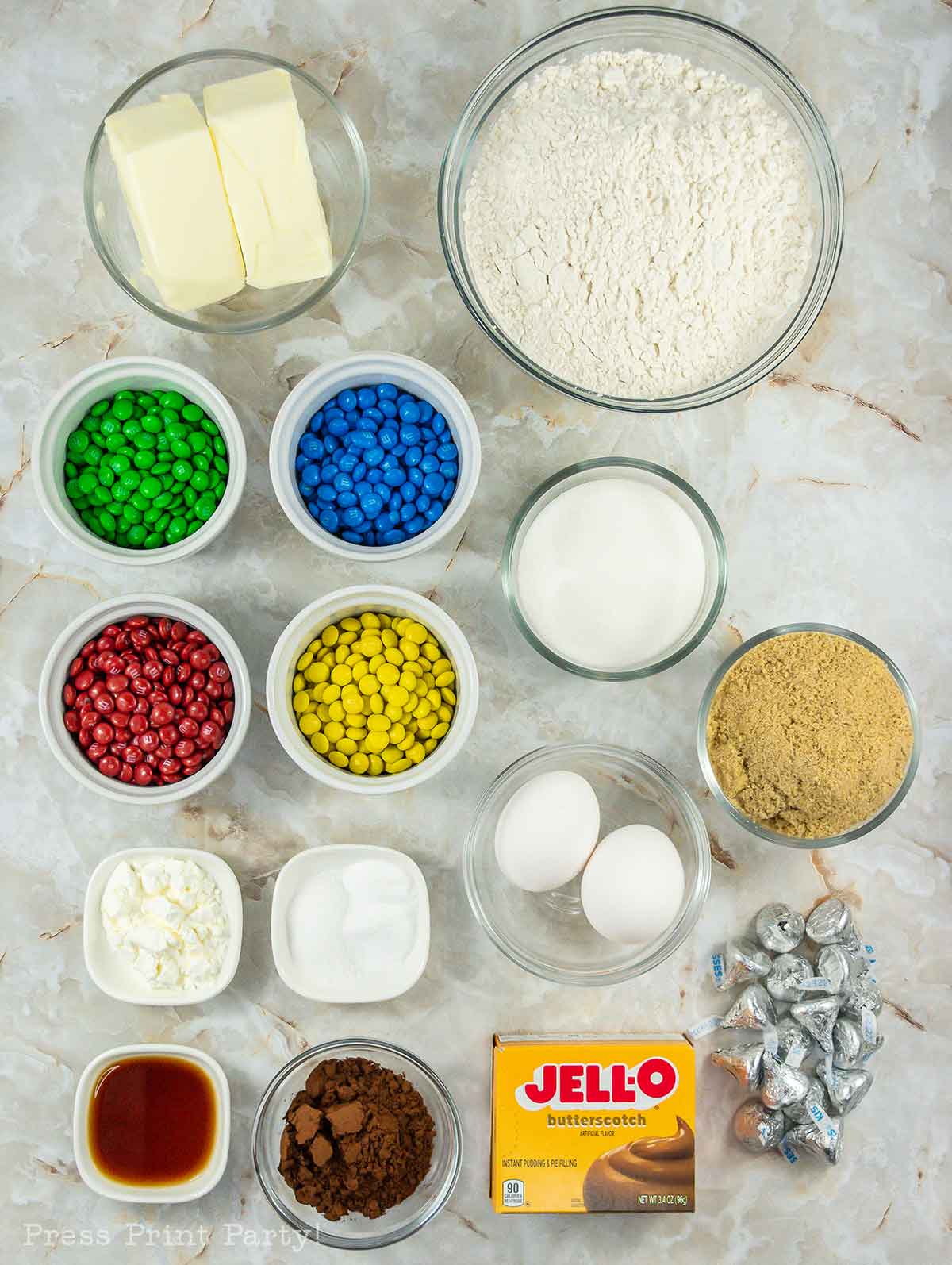 A top-down view of baking ingredients arranged on a marble surface, including flour, butter, sugar, brown sugar, eggs, cocoa powder, cornstarch, vanilla, baking soda, butterscotch pudding mix, Hershey’s Kisses, and colorful M&Ms in small bowls.