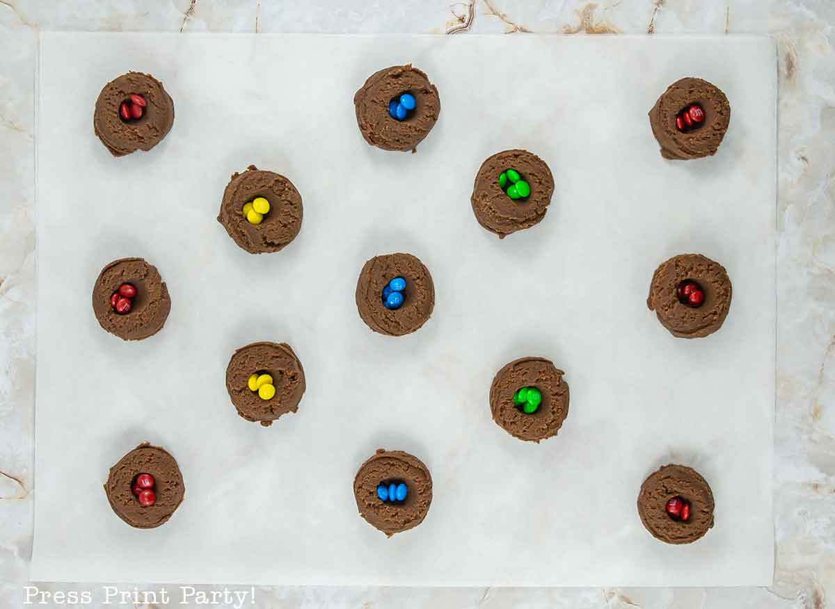 Fourteen chocolate cookie dough scoops, each topped with two colorful candy-coated chocolates, are arranged evenly on a sheet of parchment paper on a marble surface.