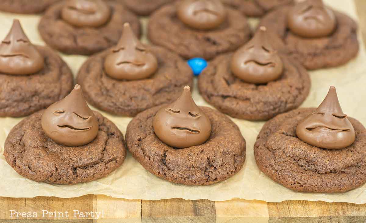 Chocolate cookies topped with chocolate drops decorated with playful, sad, or silly faces. One blue candy is visible among the cookies, which are arranged on parchment paper.