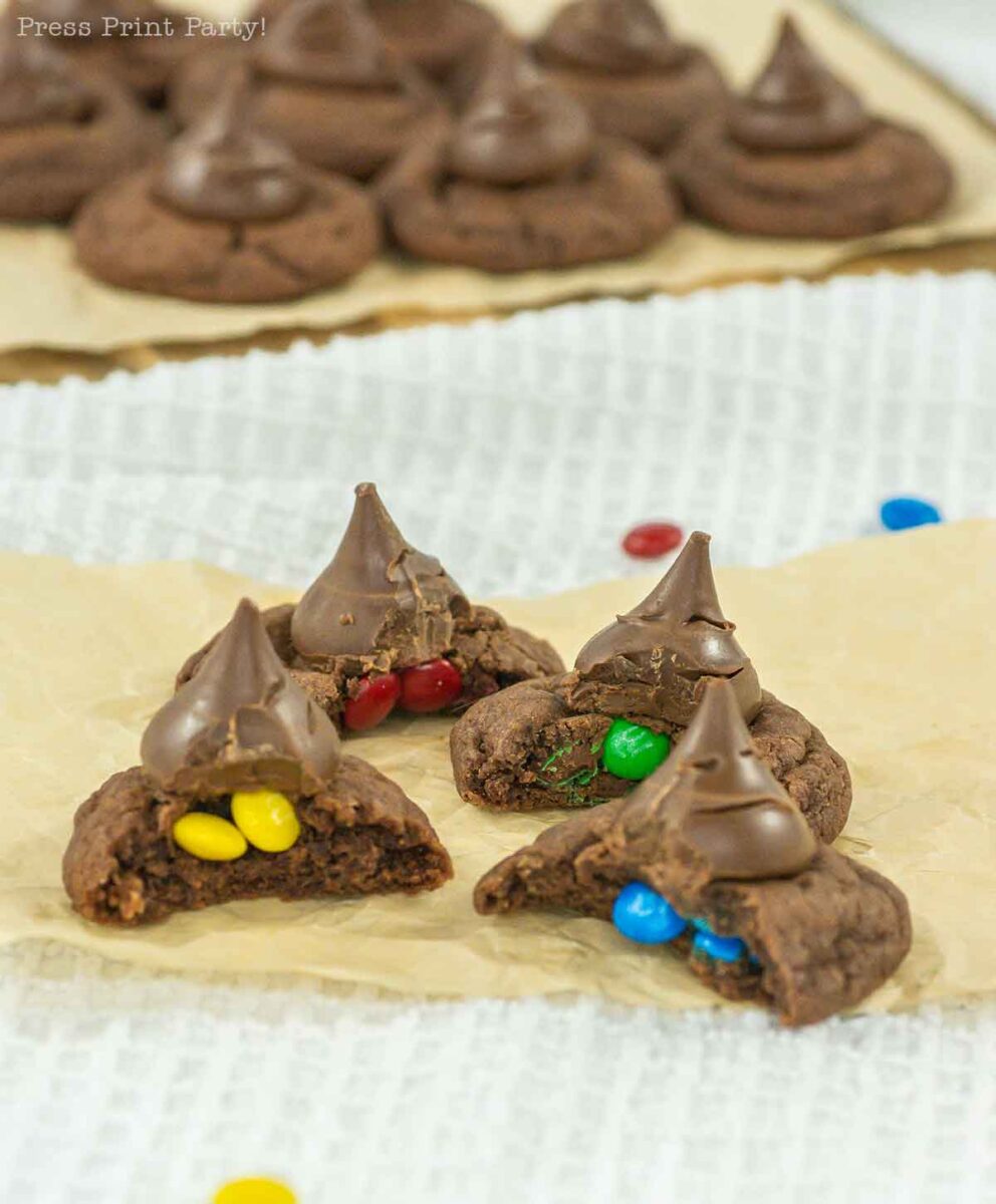 Chocolate cookies topped with a chocolate kiss, some broken open to reveal colorful candy-coated chocolates inside. More cookies are visible in the background on parchment paper.