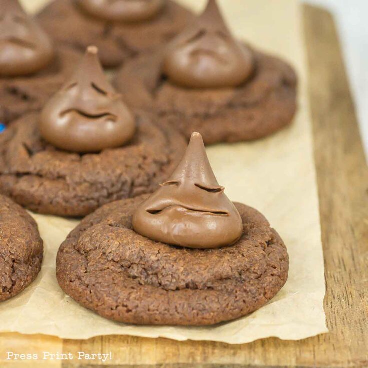 A close-up of chocolate cookies topped with chocolate kisses, with one kiss shaped to look like a smiling face. The cookies are arranged on parchment paper on a wooden board.