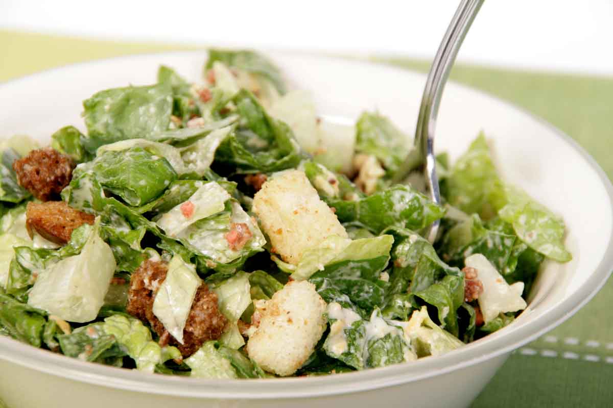 A close-up of a Caesar salad in a white bowl. The salad features crisp romaine lettuce, crunchy croutons, grated cheese, and bits of bacon, all lightly coated in creamy dressing. A fork is placed in the salad, ready to eat.