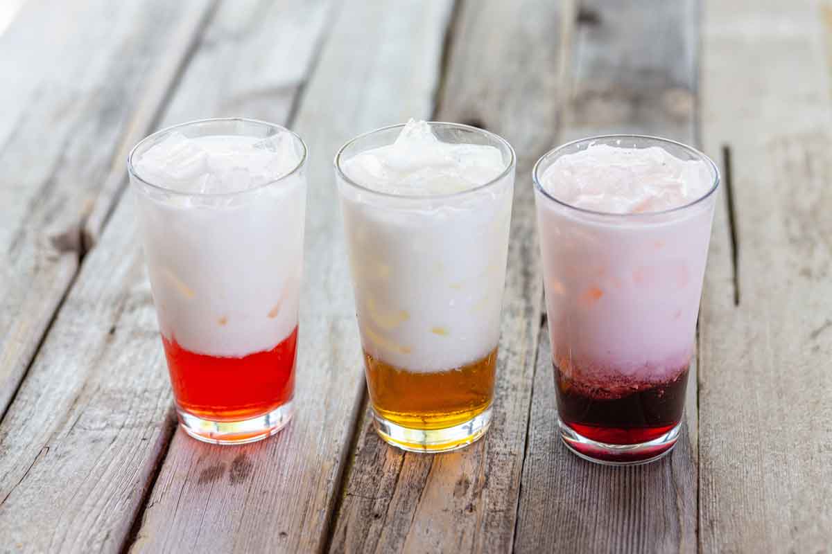 Three glasses filled with layered drinks sit on a rustic wooden table. Each glass has a white foamy top, with red, orange, and purple liquid layers at the bottom, from left to right.