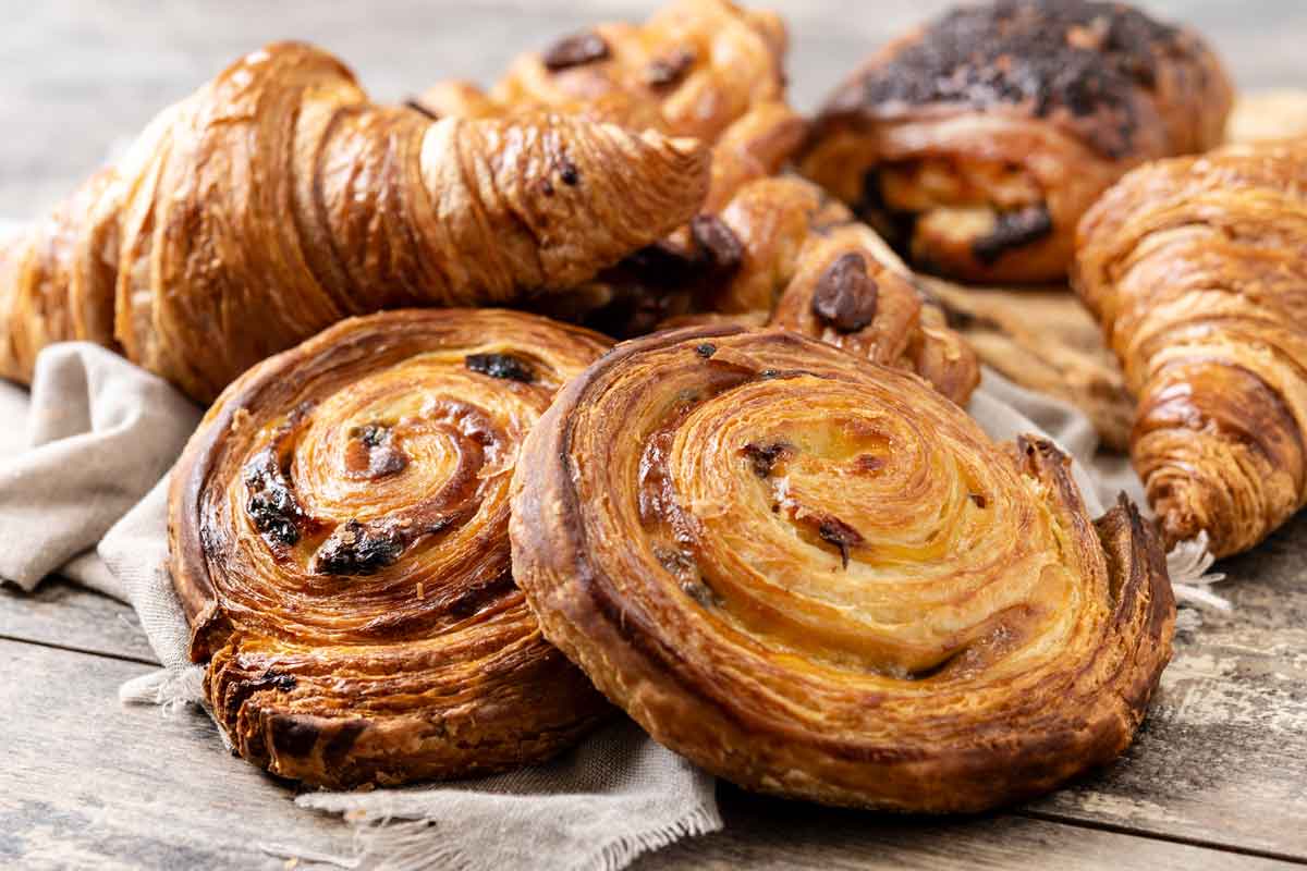 Assorted pastries on a wooden surface, including golden-brown croissants and spiraled pastries with a glossy finish and scattered raisins. A linen cloth is partially visible under the pastries.
