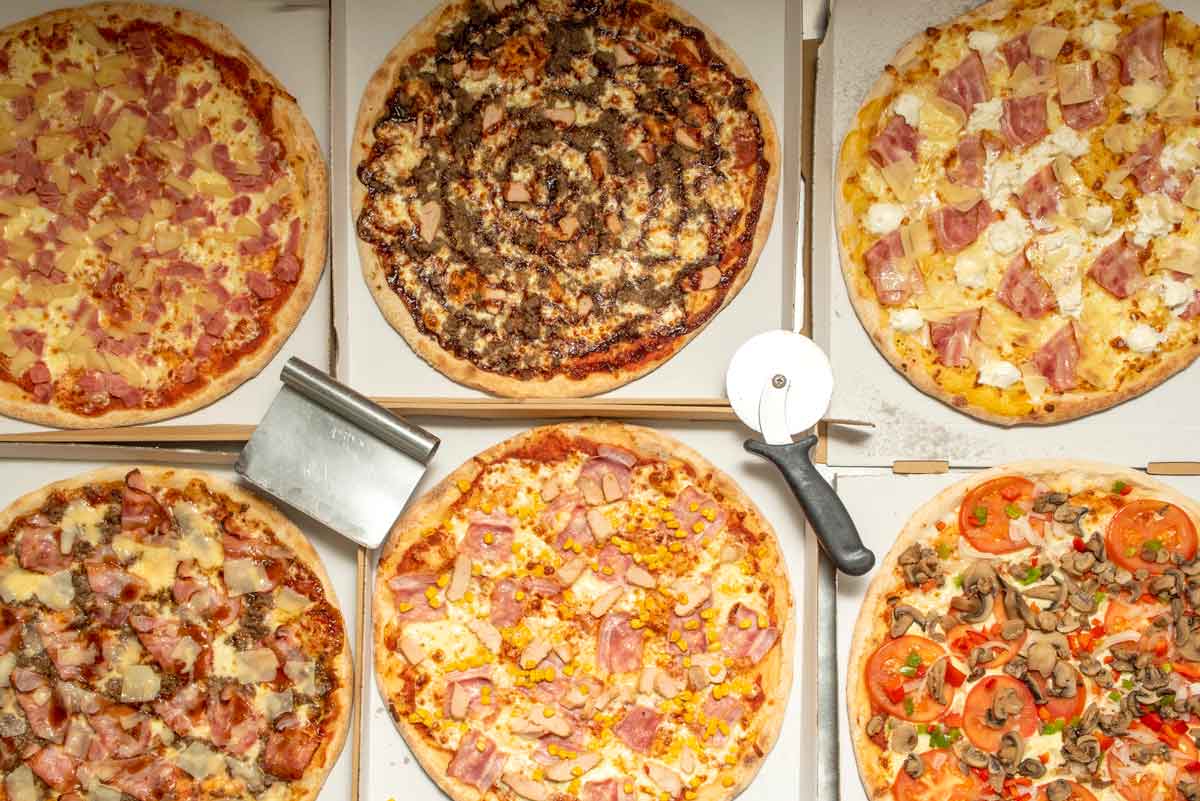 An assortment of six pizzas with different toppings, including meats, vegetables, and cheeses, are displayed in open boxes. A pizza cutter and a dough scraper are placed among the pizzas.