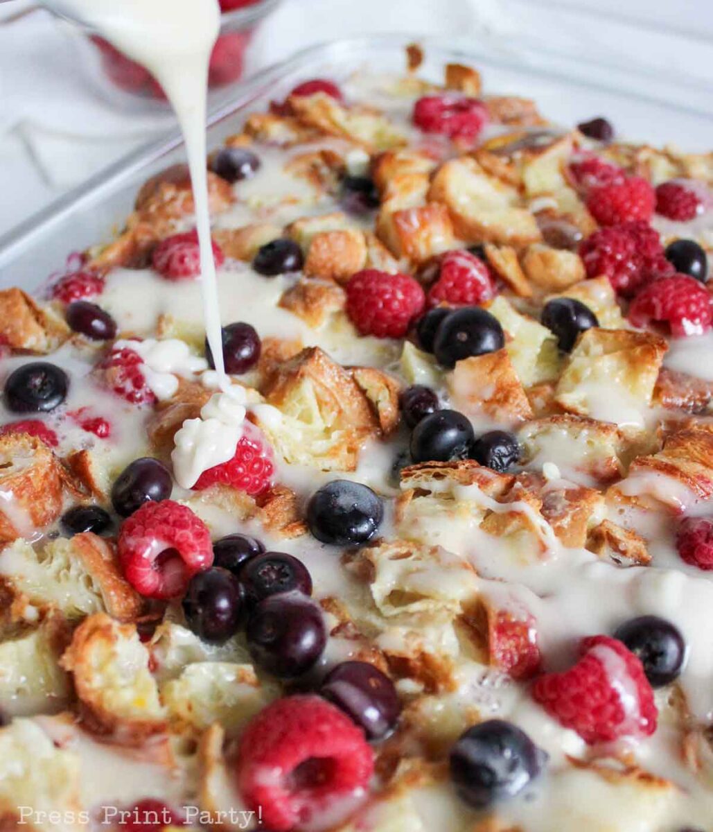 Close-up of a baked bread pudding topped with raspberries and blueberries, as a creamy white glaze is poured over it.