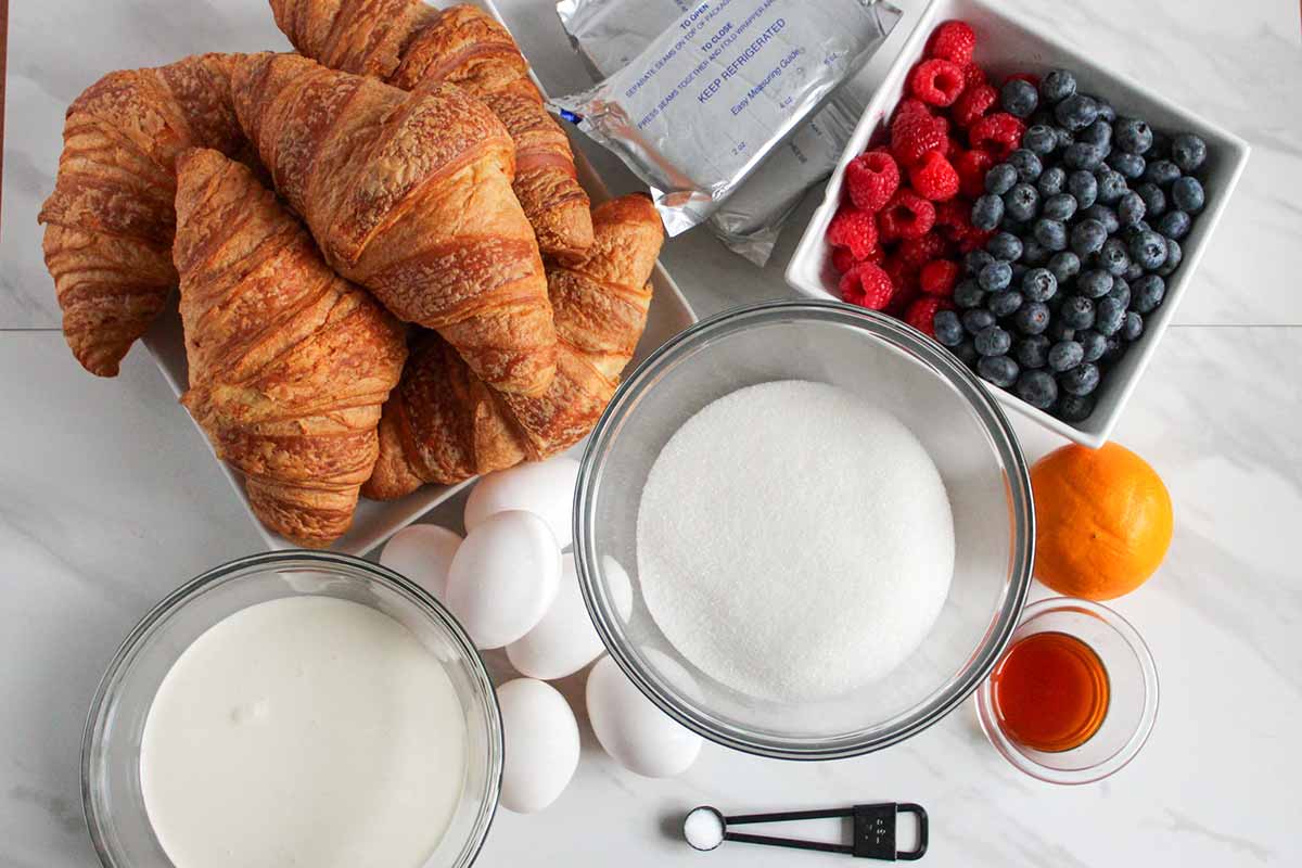 An assortment of ingredients on a countertop: croissants, cream cheese packets, a bowl of raspberries and blueberries, a bowl of sugar, eggs, cream, an orange, vanilla extract, and a measuring spoon with salt.