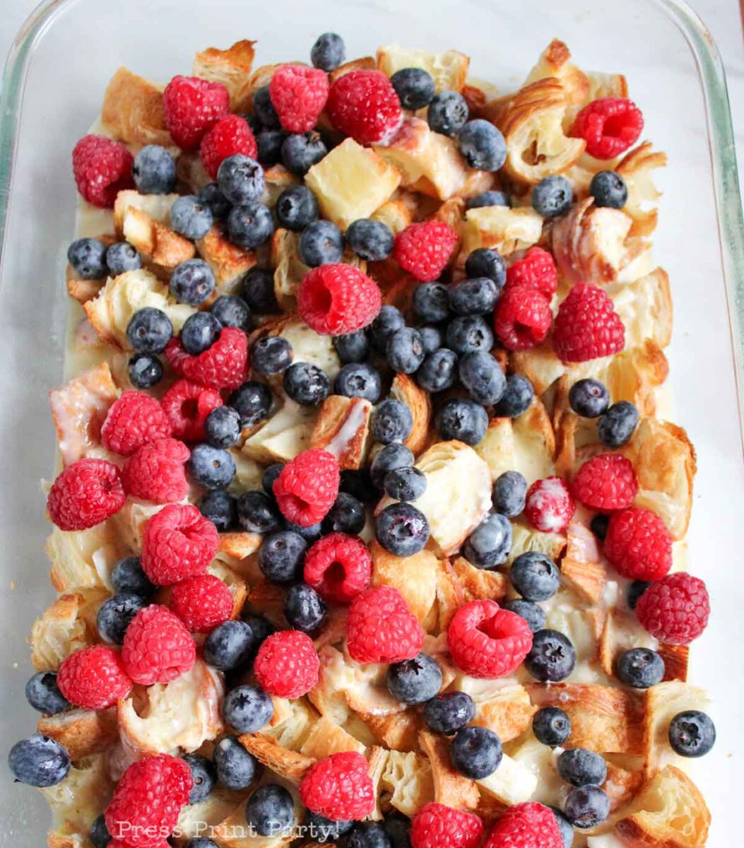 A close-up of a baked dish made with pieces of croissant topped with fresh raspberries and blueberries in a glass baking dish.