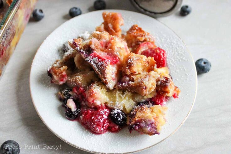 A plate of baked French toast casserole topped with powdered sugar, featuring visible pieces of bread, raspberries, and blueberries, with more berries scattered around on the table.