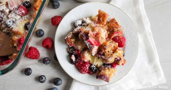 A plate of bread pudding topped with fresh raspberries and blueberries, dusted with powdered sugar, sits on a white napkin next to a baking dish and scattered berries.
