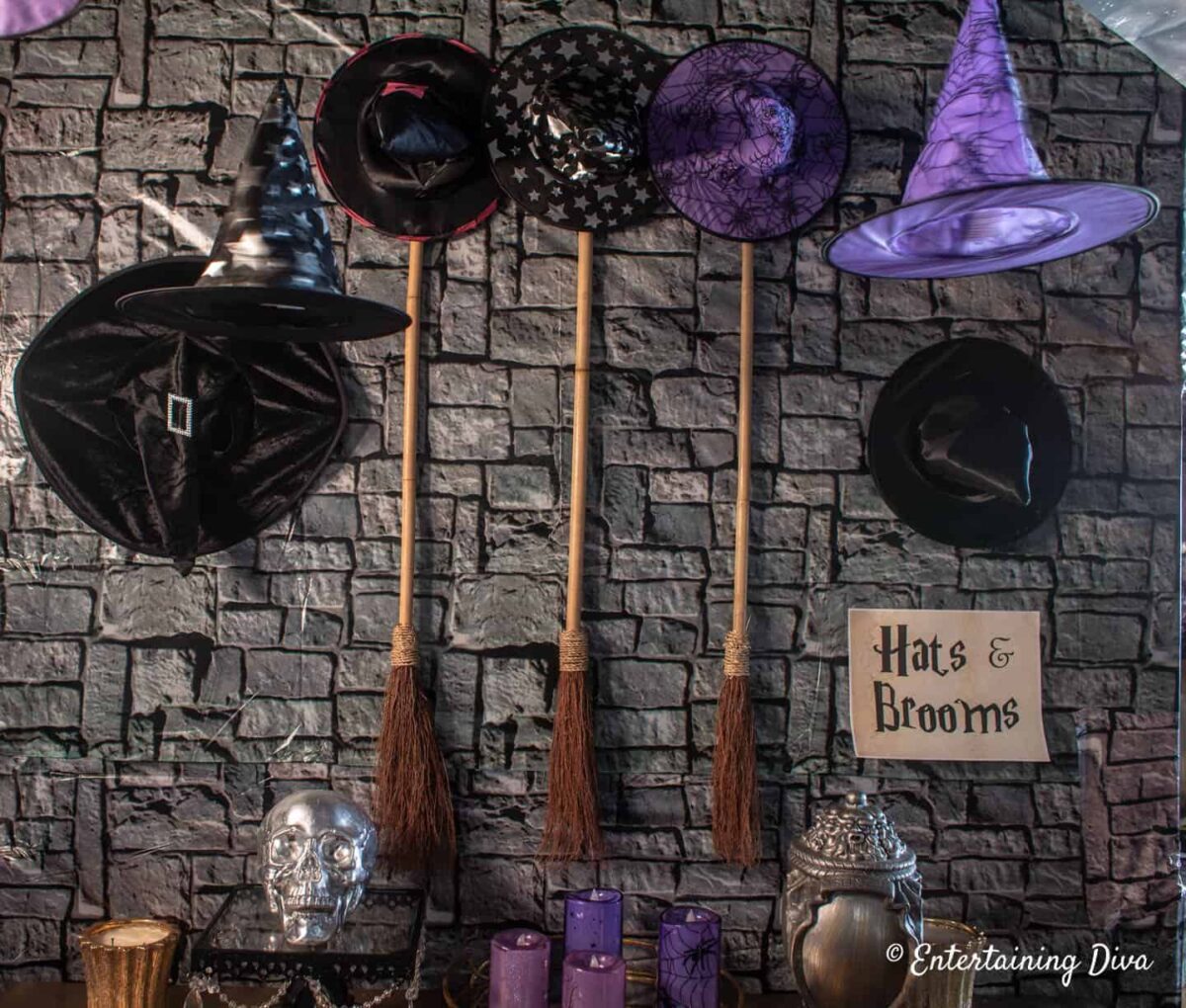 Four witch hats and two brooms hang on a gray stone wall with a sign reading "Hats & Brooms." Perfect for Harry Potter birthday parties, the display features purple candles, a silver skull, and jars for a magical Halloween vibe.