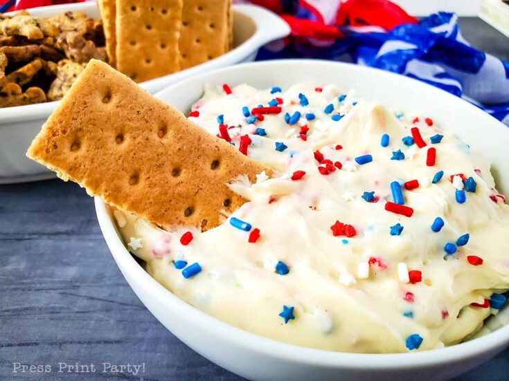 A white bowl filled with red white and blue dip, topped with festive sprinkles and garnished with a graham cracker. In the background, more graham crackers and patriotic ribbons complete the scene.