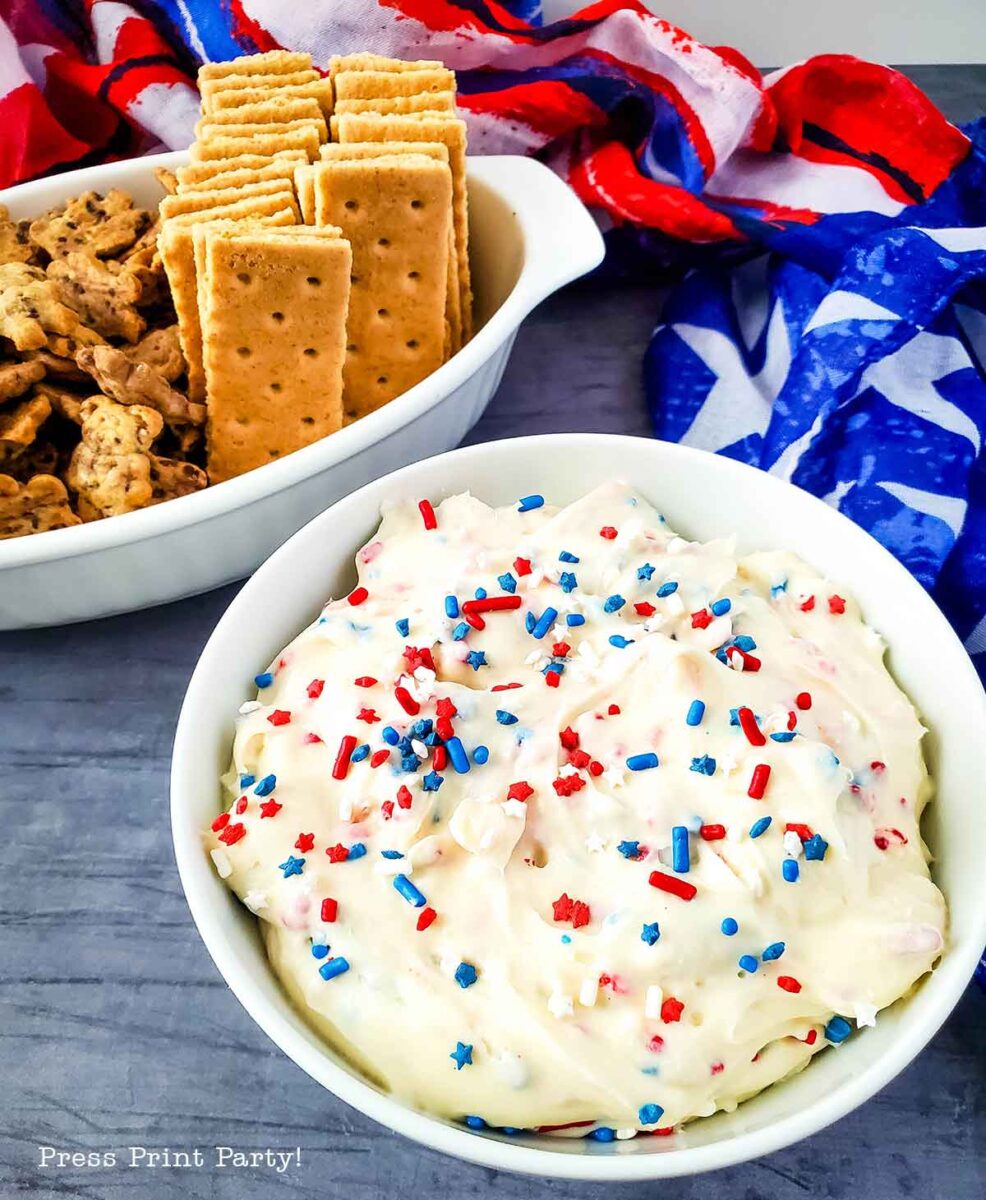 A bowl of creamy red white and blue dip topped with festive sprinkles sits next to a dish of graham crackers and animal cookies. A vibrant red, white, and blue decorative cloth is in the background.