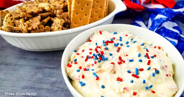 A festive red white and blue dip sits in a bowl, topped with colorful sprinkles beside graham crackers and pretzel crisps on a gray surface, with a patriotic-themed cloth in the background.