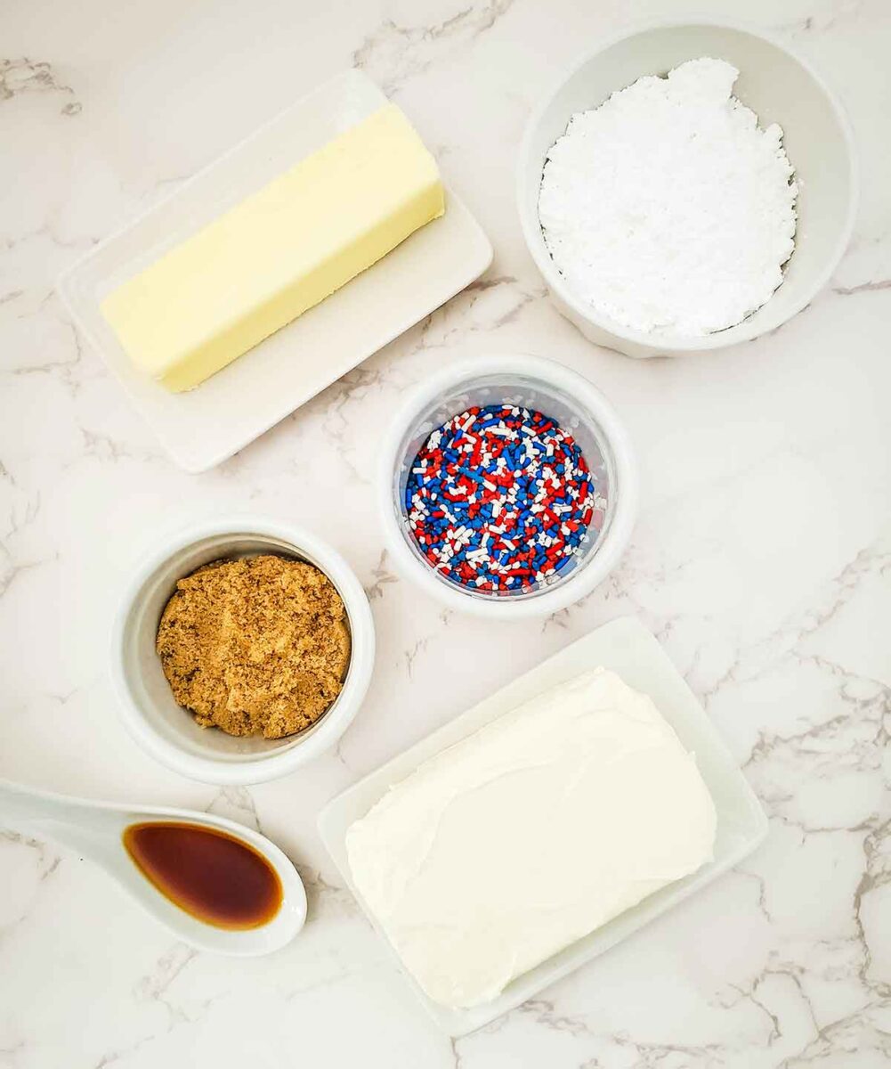 Top view of baking ingredients on marble: a stick of butter, bowls of powdered sugar, brown sugar, red white and blue sprinkles—perfect for creating a festive red white and blue dip—plus cream cheese and a spoon with vanilla extract.